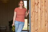 Frau in ockerfarbenem Tshirt und Jeans am Eingang eines Hauses mit Holzfassade.