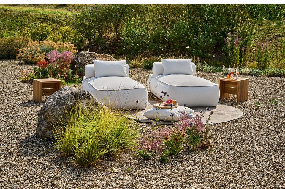 Zwei graue Loungesessel stehen auf Schotter in einem Garten mit Teppich und Beistelltischen aus Holz, drumherum einige Pflanzen