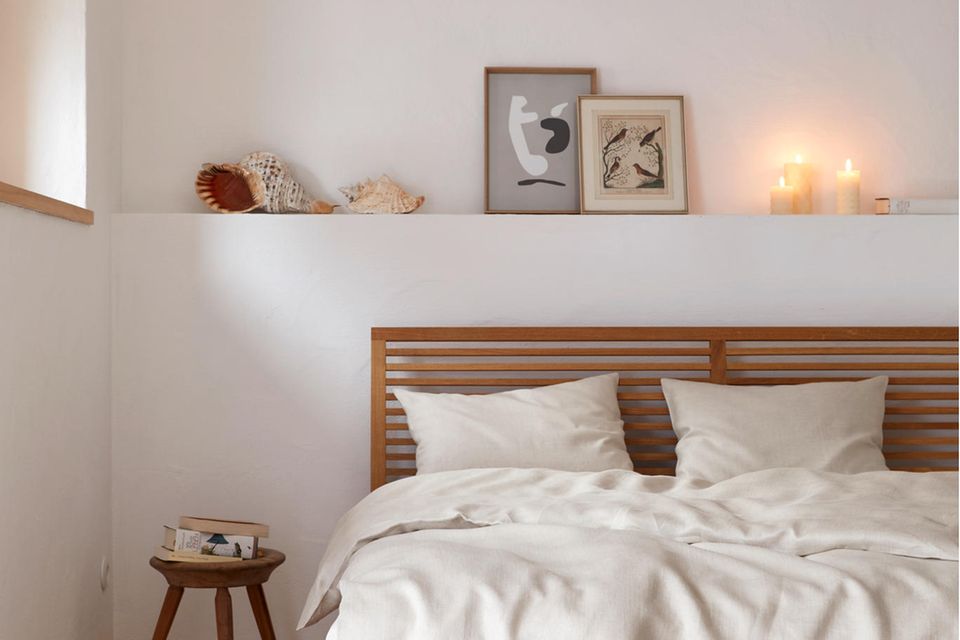 Holzbett mit heller Bettwäsche steht vor einer weißen Wand, darüber Bilder, Deko und Kerzen