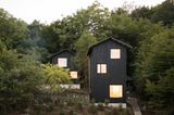 Schwarze Holzhäuser mit Satteldach mit beleuchteten Fenstern inmitten von hohen Bäumen