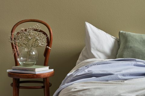 Bett vor einer grünen wand, daneben ein Holzstuhl mit Büchern und einer Vase und darüber eine Leuchte