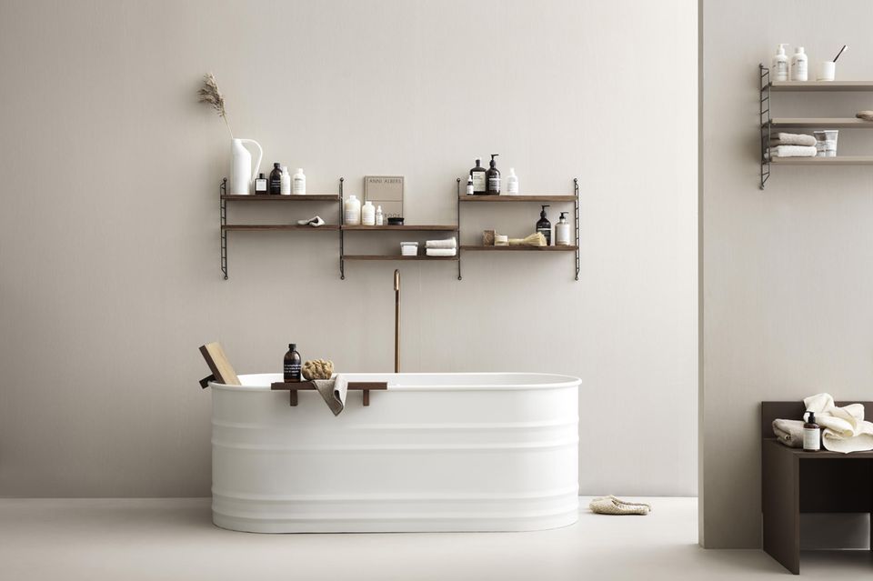 Badezimmer mit weißer Badewanne vor einer hellen Wand, dahinter ein Wandregal mit Deko
