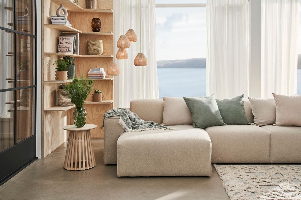 Wohnzimmer mit beiger Couch, grünen Kissen und Holzmöbel, dahinter eine große Fensterwand