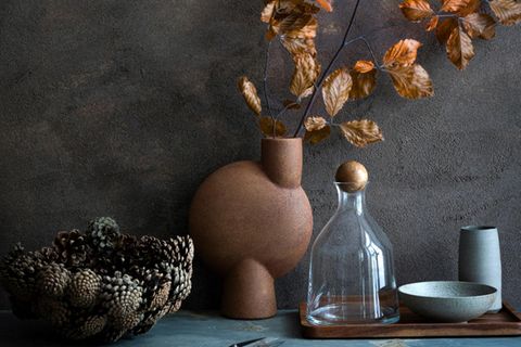 Braune Vase mit Ast mit Herbstlaub steht vor einer dunkelgrauen Wand, daneben ein Tablett mit Schalen und einer Glasflasche