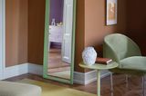 Grüner Spiegel lehnt an terrakottafarbener Wand, davor ein gelber Teppich und hinten ein grüner Sessel
