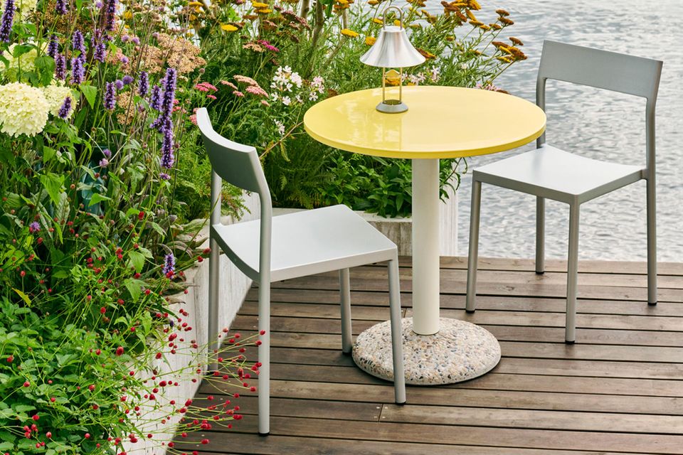 Gartentisch mit gelber Platte auf einer Holzterrasse neben einem blühenden Beet, dahinter Wasser
