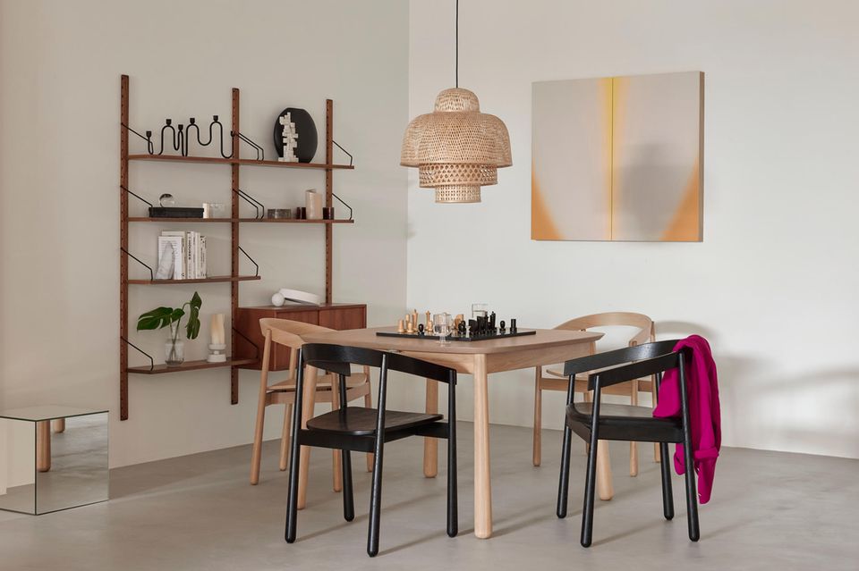 Esszimmer mit Holztisch und vier Stühlen, darauf ein Schachbrett, dahinter ein Holzregal mit Deko und ein Bild an der Wand