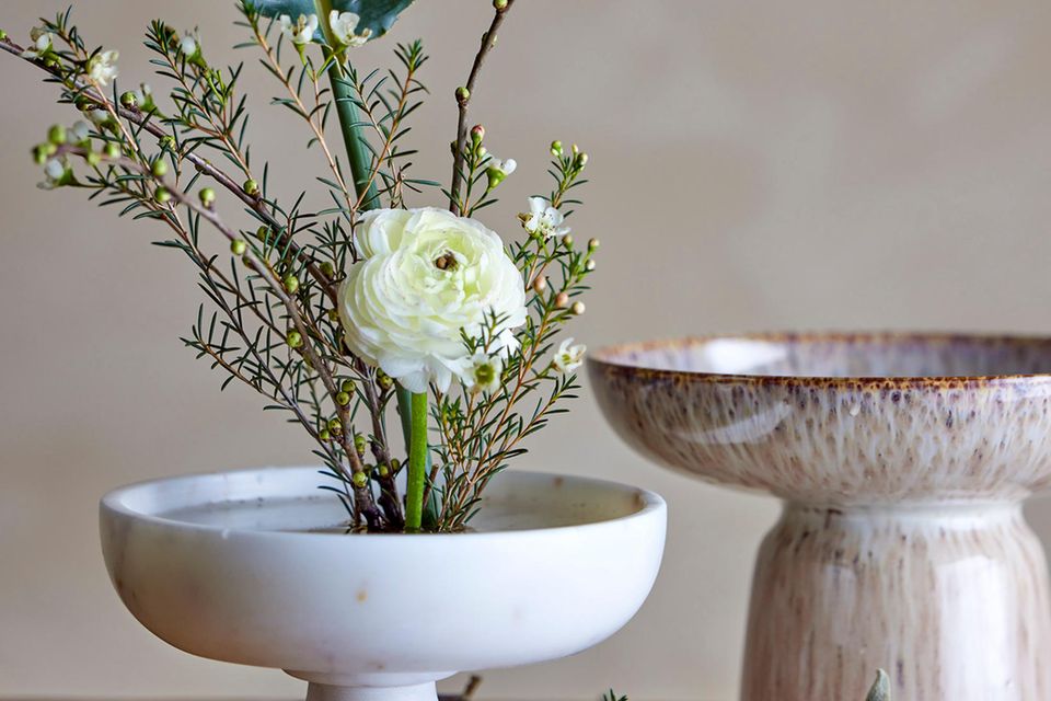 Weiße Schale aus Marmor mit Blumen udn pflanzen auf einem Holztisch, dahinter eine weitere Schale mit Fuß