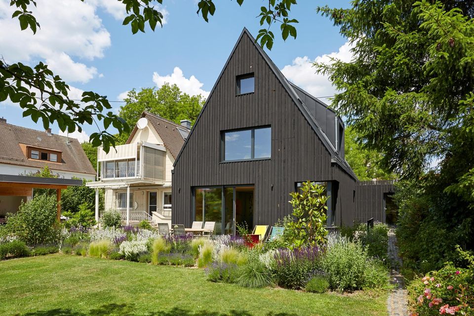 Garten mit schwarzem Holzhaus und spitzem Dach, dahinter ein helles Haus
