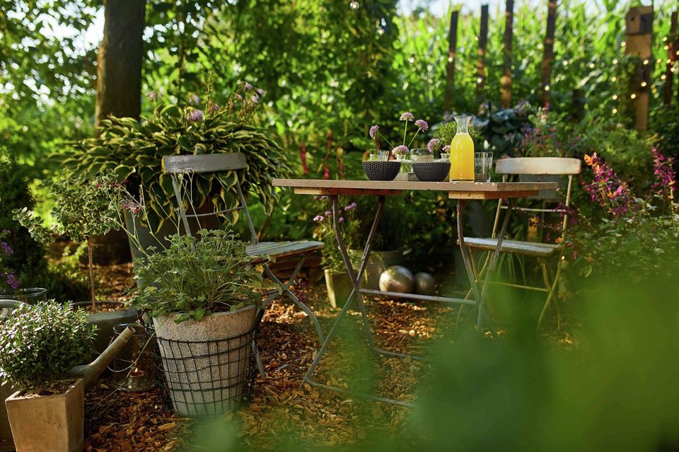 Gartenecke mit Tisch und Stühlen und Rindenmulch auf dem Boden, daneben Topfpflanzen